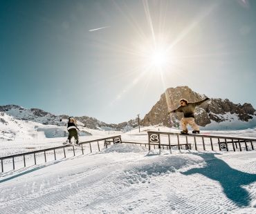 Two snowboarders boardsliding rails